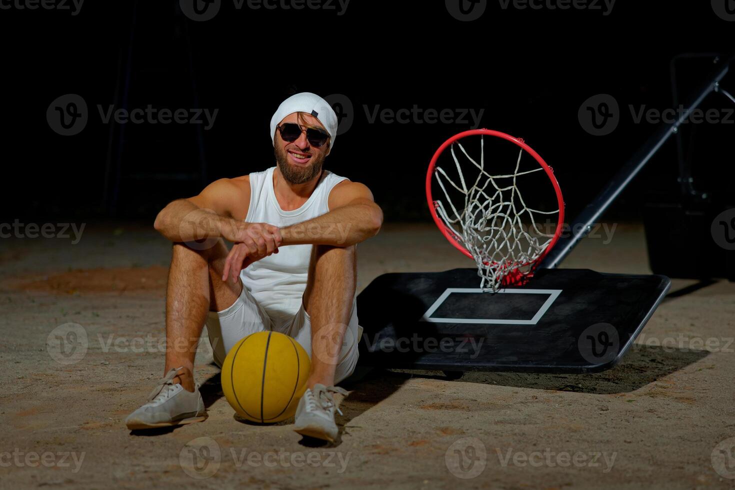 Basketball player sits on the court wearing sunglasses, relaxed after an intense practice session at dusk. photo