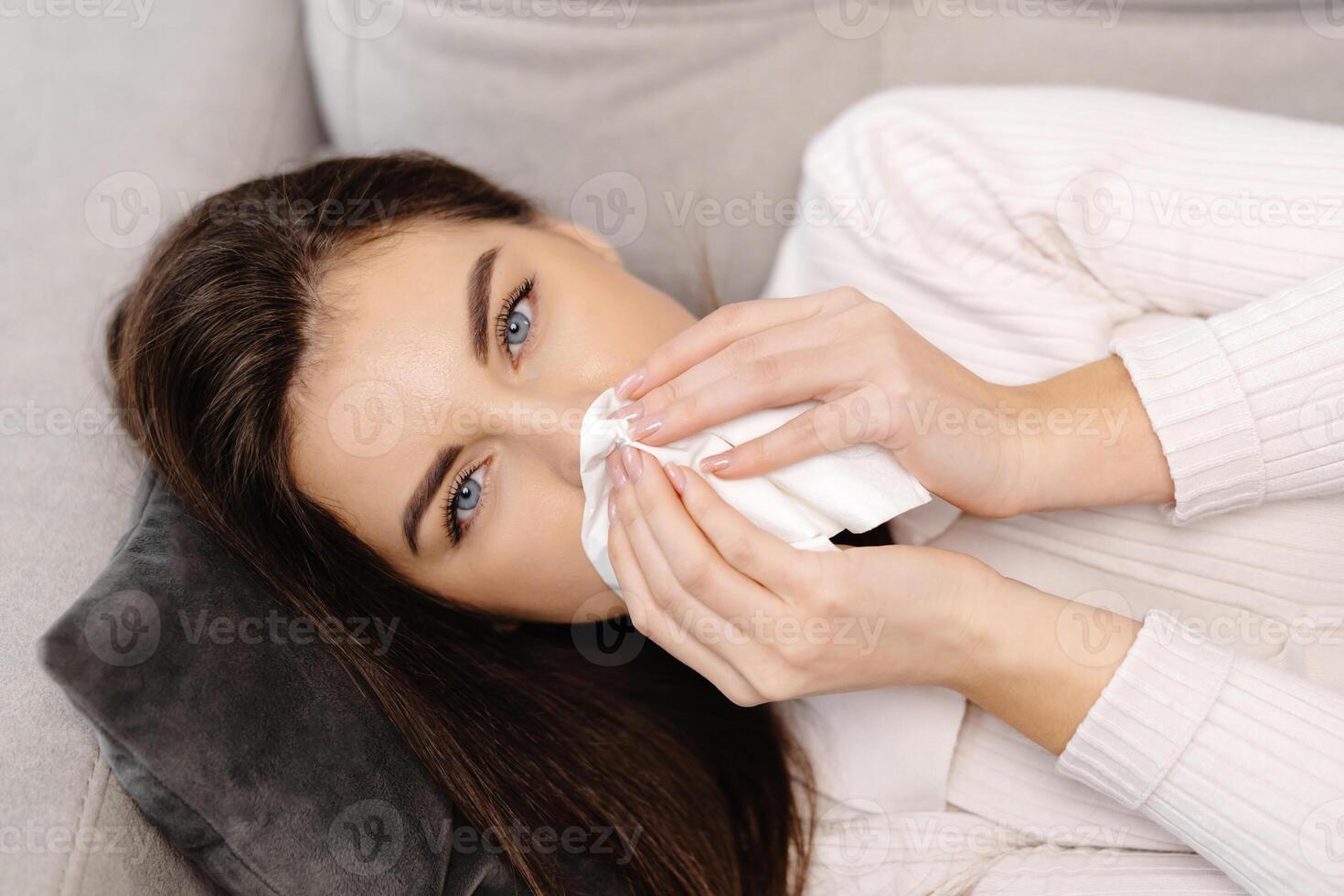 Unhappy sad young female with plaid, suffering from fever and flu on sofa, blowing nose in napkin in living room interior photo