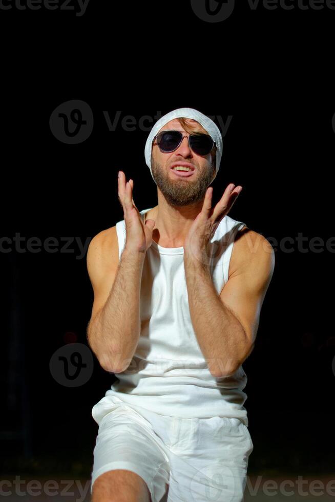 Man wearing a tank top, sunglasses, and a beanie poses confidently at night against a dark background photo