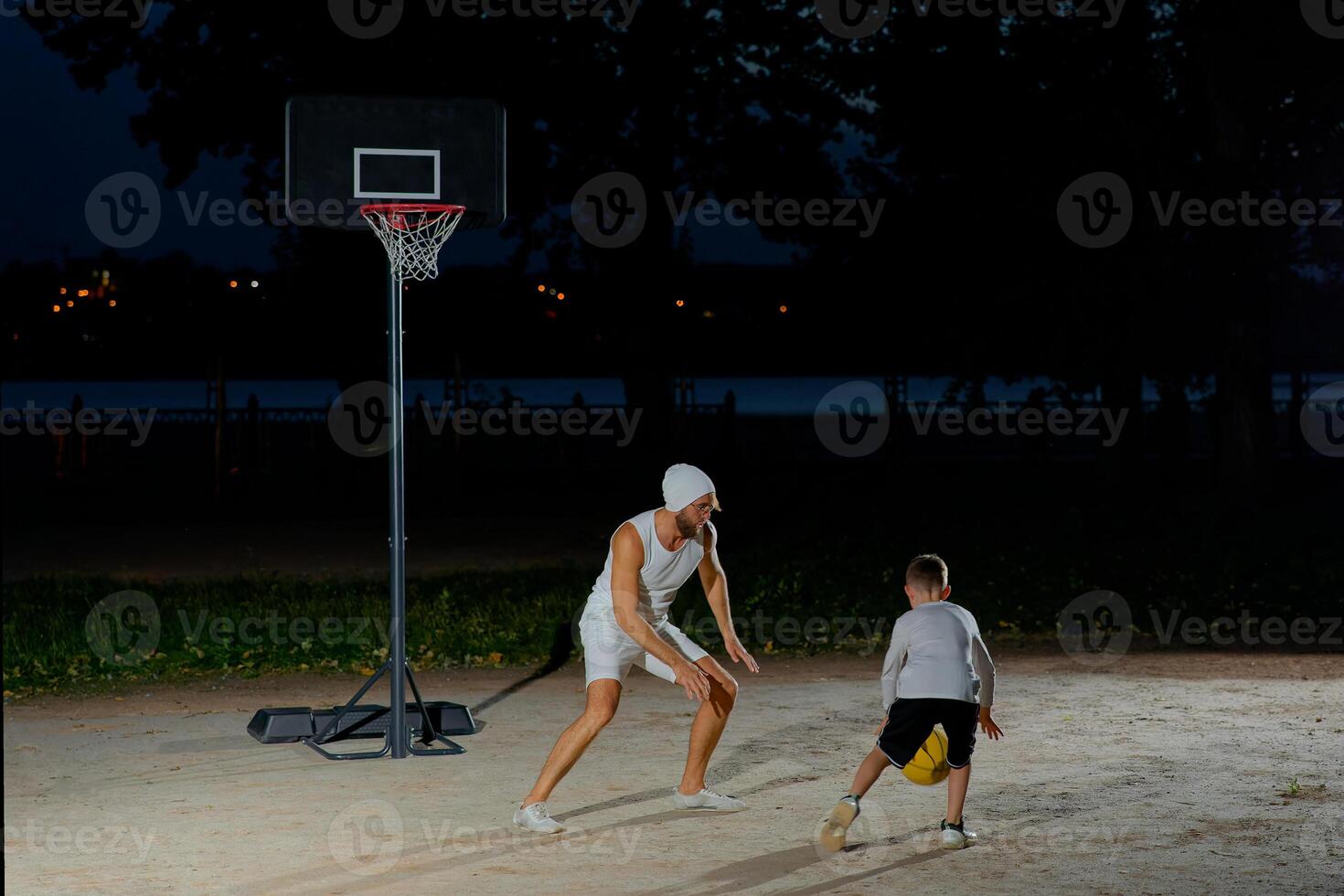 A father and son enjoy a night basketball game at a local park under bright lights focusing on teamwork photo