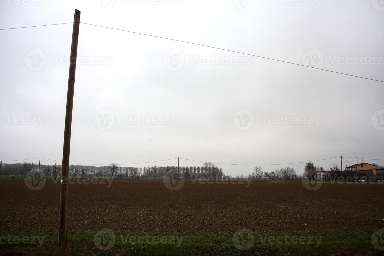 Ploughed field with a grove and buildings in the distance framed by a over head cable and a wooden pole on a cloudy day in the italian countryside photo