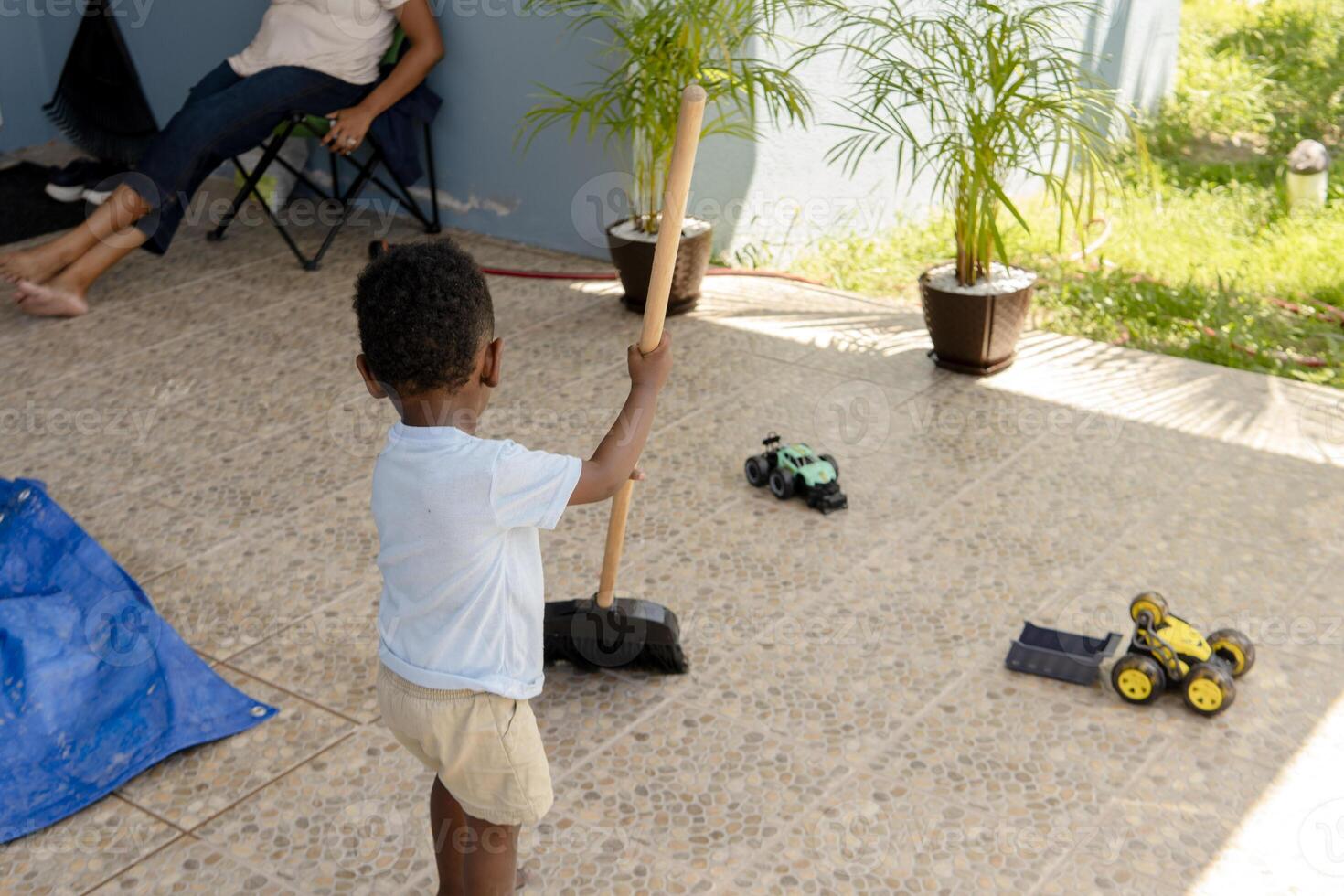 Little boy sweeping patio while mother relaxing and watching him play photo