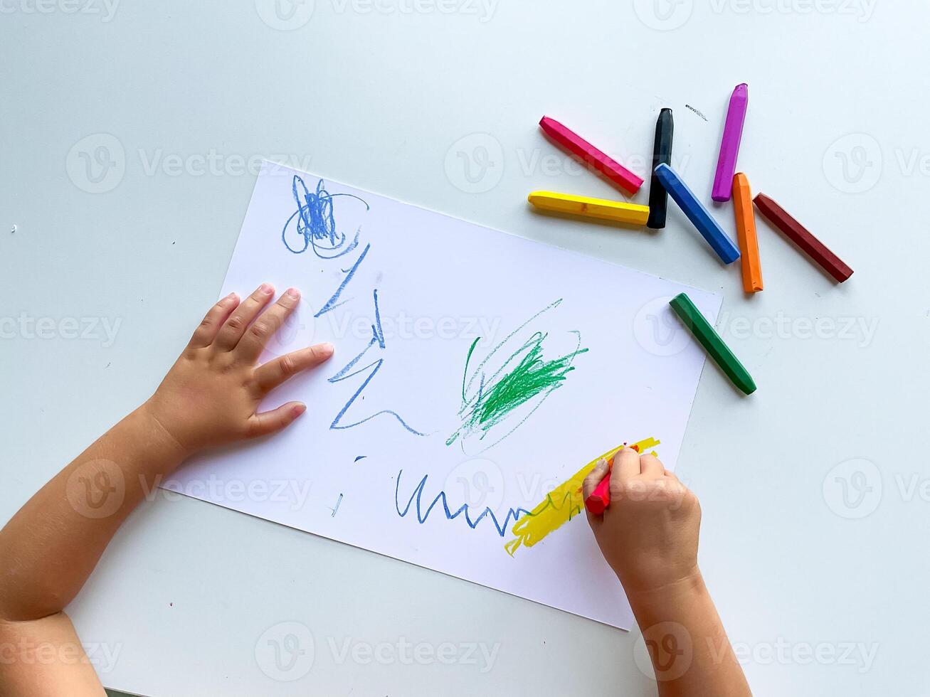 Child drawing with multicolored crayons on paper surface photo