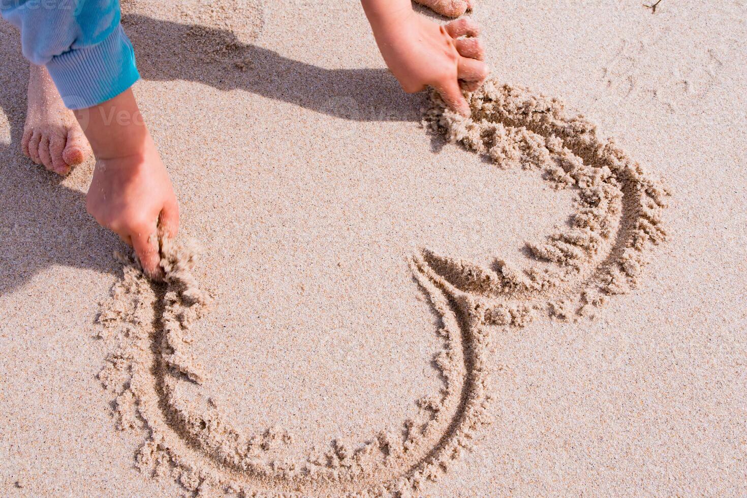 Child drawing heart shape in sand on beach photo
