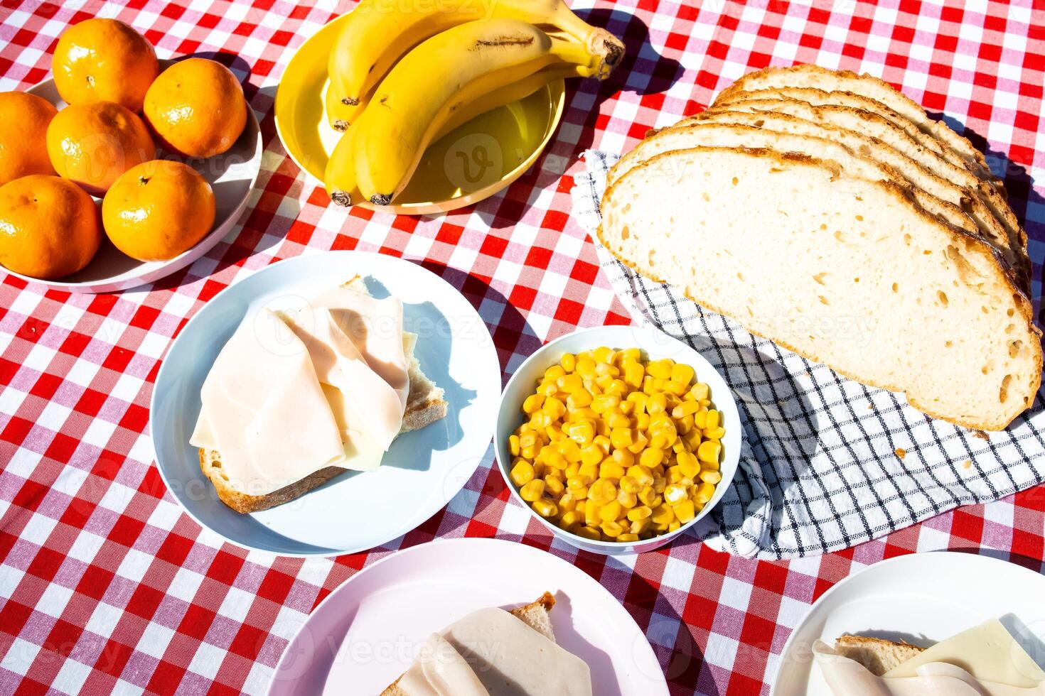 Outdoor picnic table with sliced bread, cheese, fruit, and corn on red checkered cloth photo