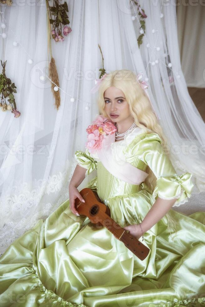 young blonde with guitar in hands in medieval costume sits on white background made of fabric. dress of soft green color with pink ribbon and pink flowers. pearls on her neck photo