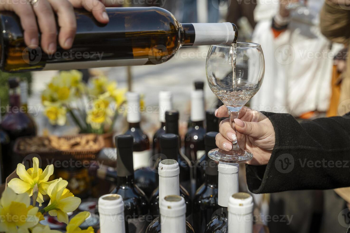 Close-up of a person's hand pouring white wine into a glass, many bottles of wine nearby. Yellow flowers in the background, shallow depth of field. The image conveys the elegance of a fine dining experience. photo