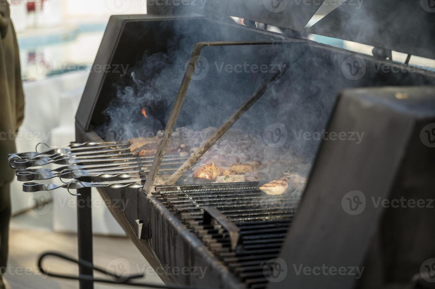 cooking meat on grill at barbecue party in summer garden. Food, people and family time concept. pork on skewers roasting on coals in large metal grill device. smoke from fire, no people photo