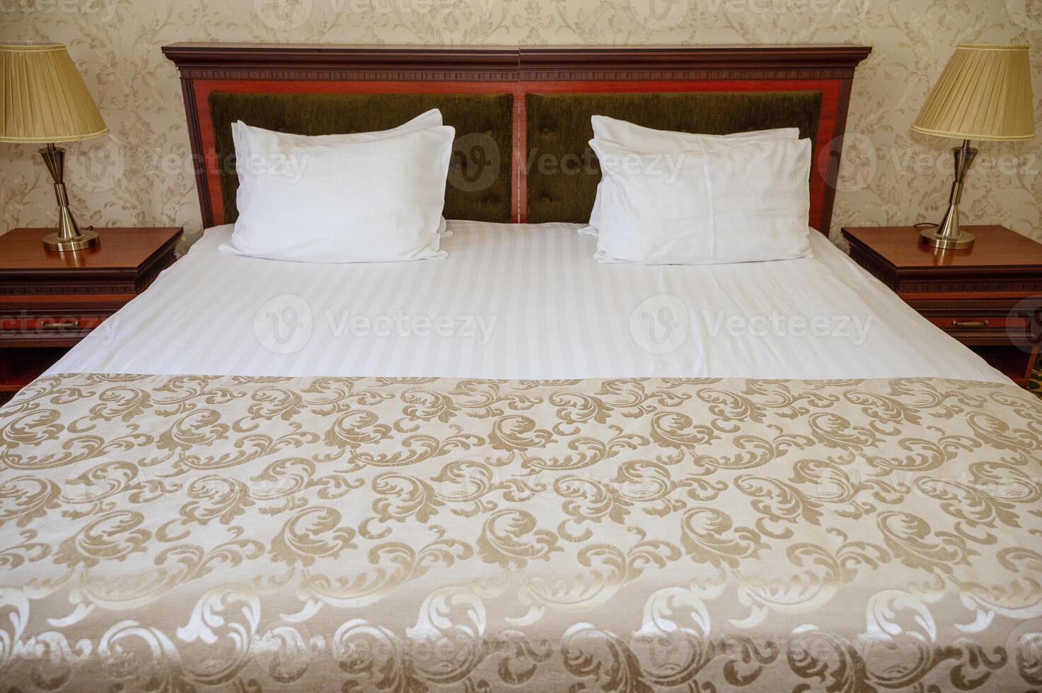 Double bed with wooden, dark red, headboard, clean white bed linen and pillows. bedspread and wallpaper of the same golden-beige shade. On the mirrored nightstands are table lamps with golden lampshades photo