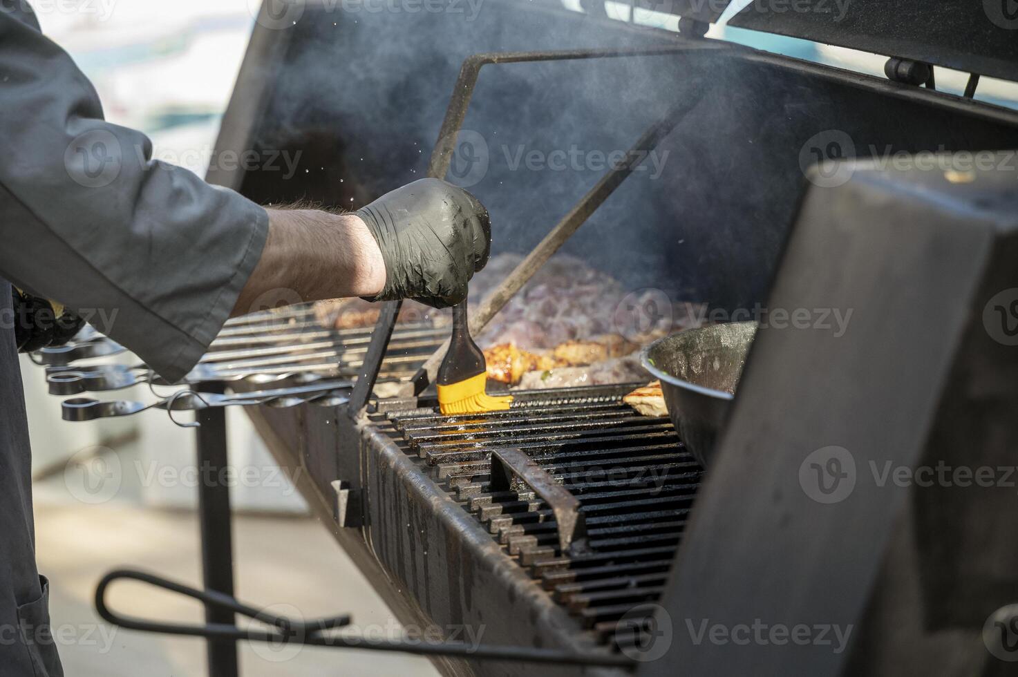 Male cook preparing meat on grill at barbecue party in summer garden, cook's hands in gloves brushing meat. Food, people and family time concept. pork on skewers roasting on coals in metal grill device photo