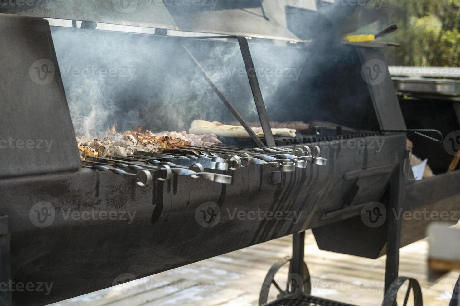 cooking meat on grill at barbecue party in summer garden in park in restaurant. Food, people and family time concept. pork on skewers roasting on coals in big metal grill device photo
