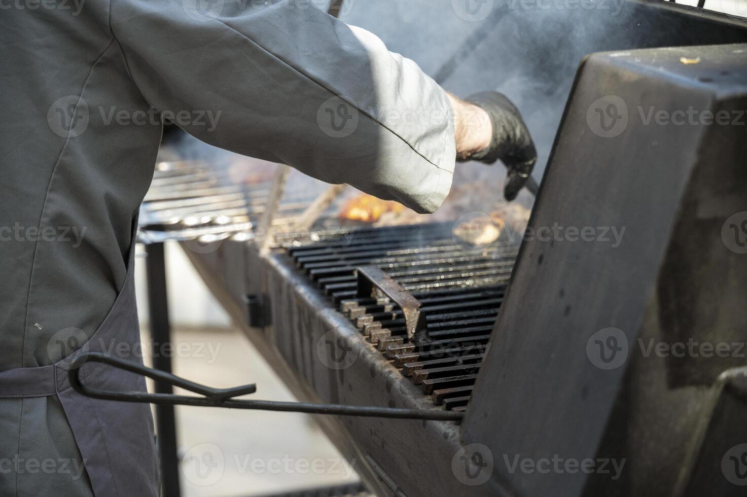 Male cook preparing meat on grill at barbecue party in summer garden,chefs in gloves and apron. Food, people and family time concept. pork on skewers roasting on coals in big metal grill device photo