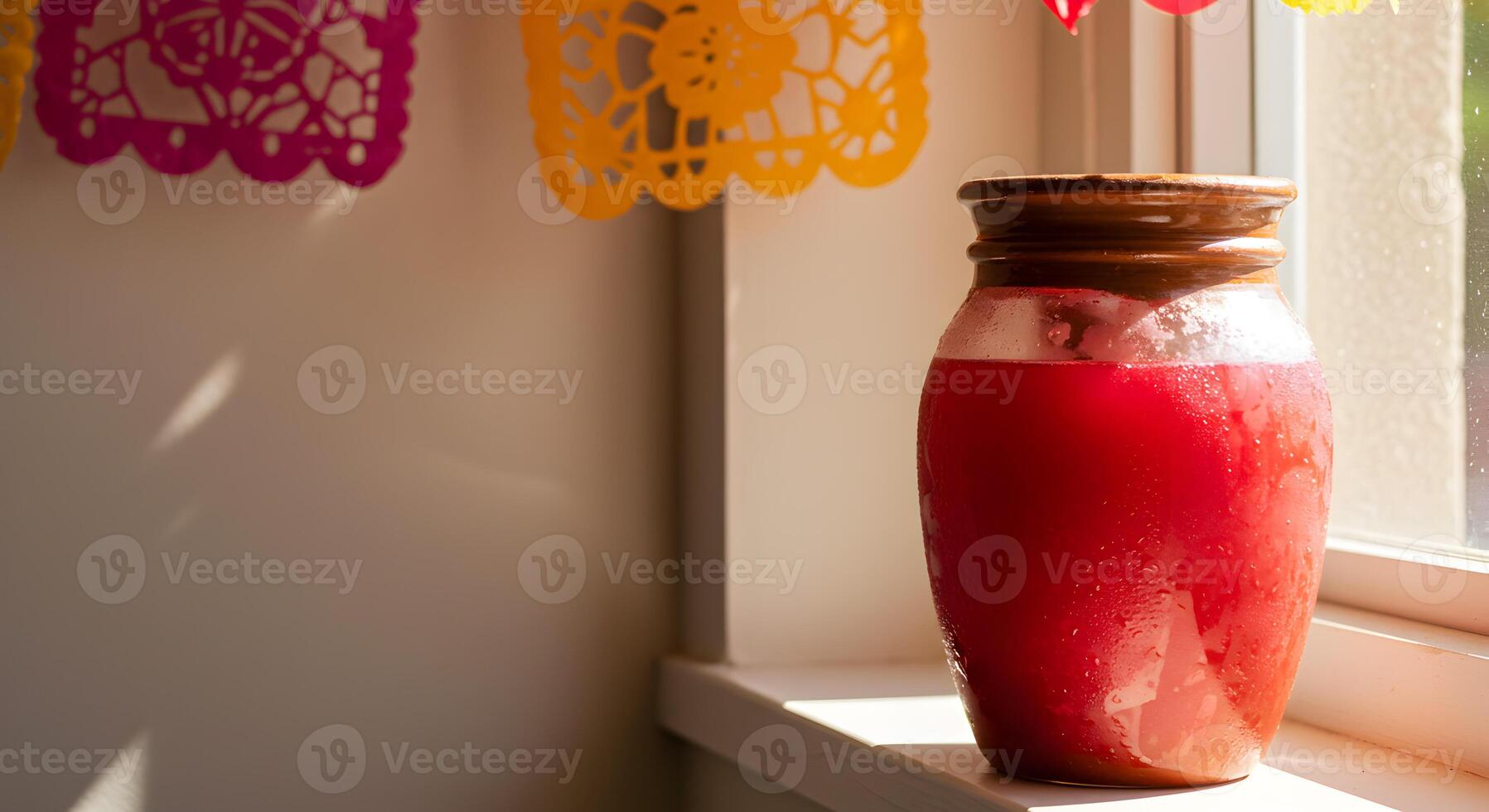 Clay jar filled with red agua fresca on sunny windowsill with papel picado decor photo
