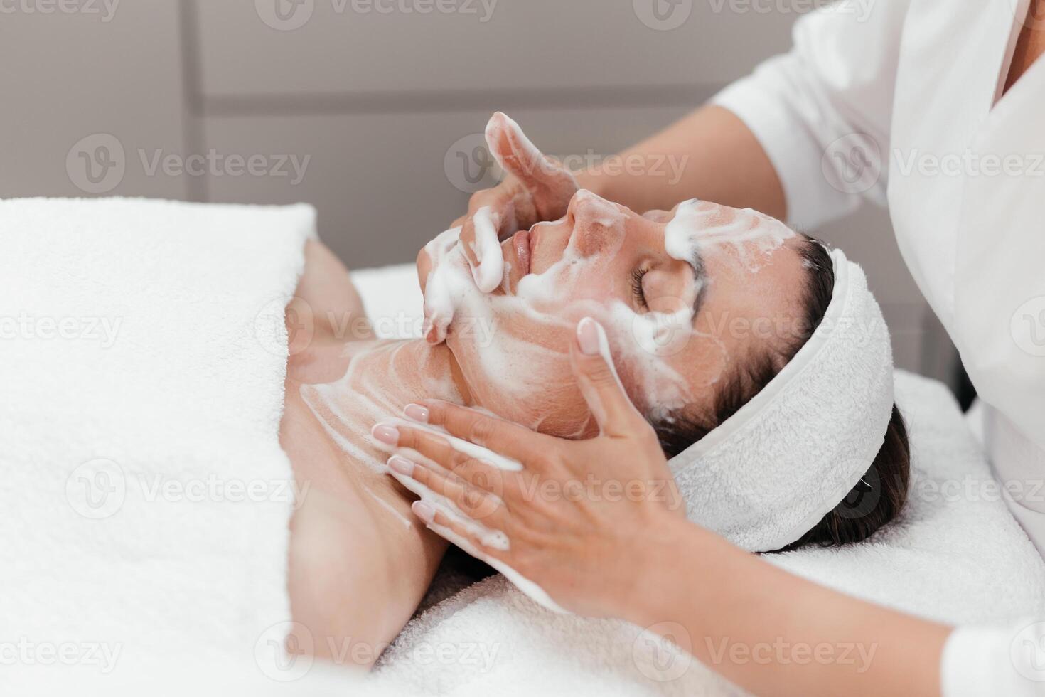 Cosmetologist doing female face massage with creamy foam, skin beauty treatment, improving appearance. Relaxed girl getting professional facial rejuvenating, cleansing, moisturizing in beauty clinic photo
