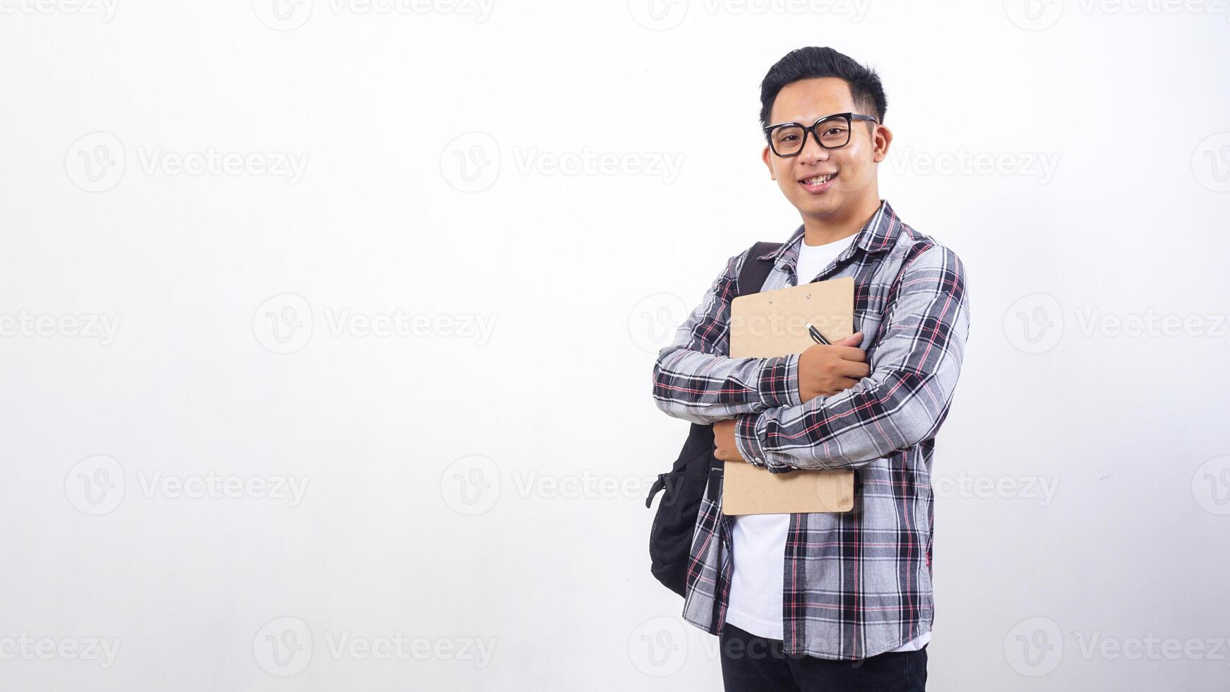 Young Asian male college student with a backpack holding wooden clipboard and pen in front his chest isolated on white background photo