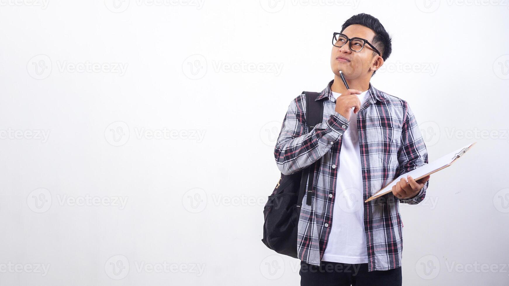 Handsome young Asian male college student with a backpack holding wooden clipboard and pen while showing gesture of thinking isolated on white background photo