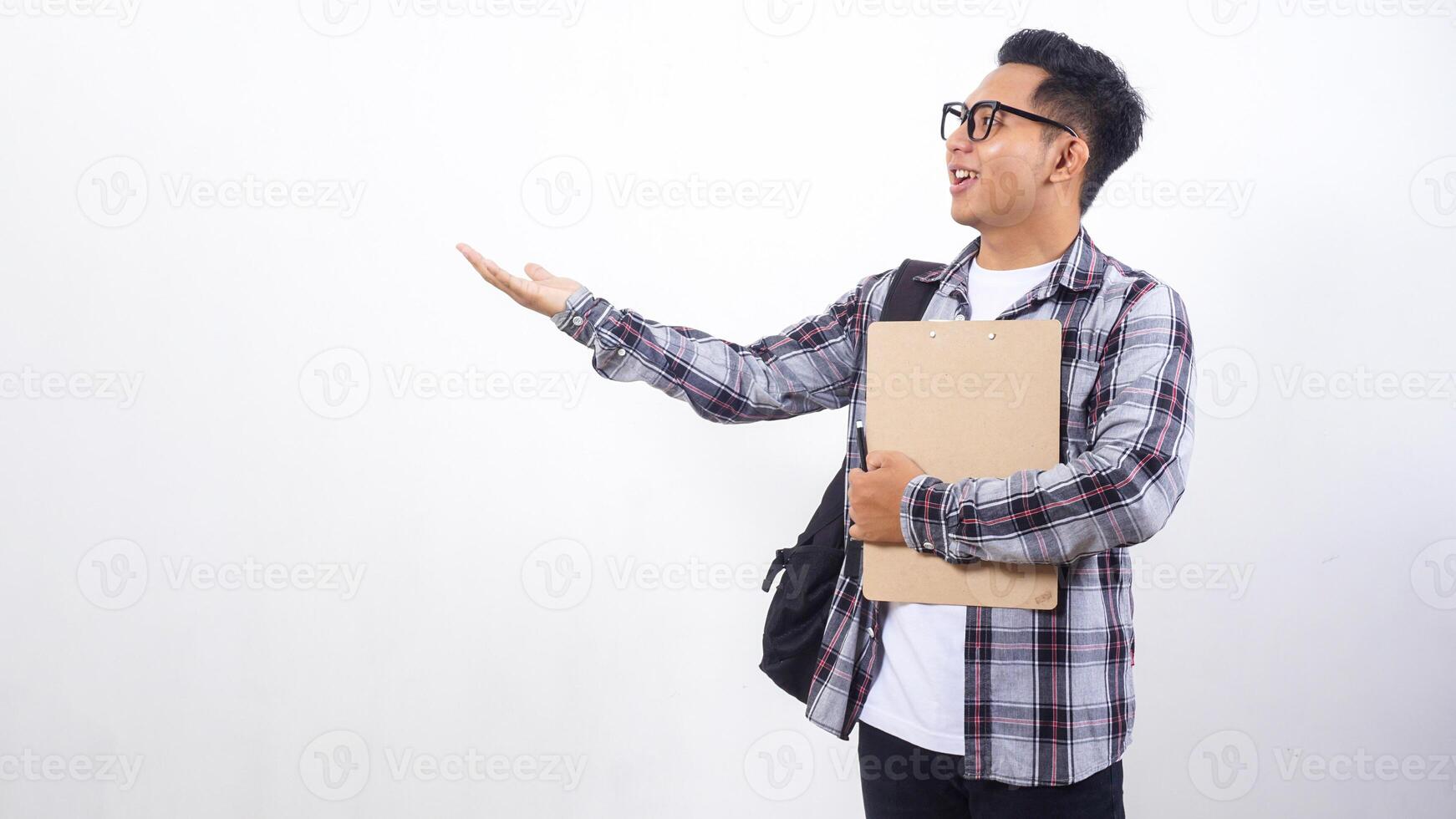 Young Asian male college student with a backpack holding wooden clipboard and pen while presenting empty space with his hand isolated on white background photo