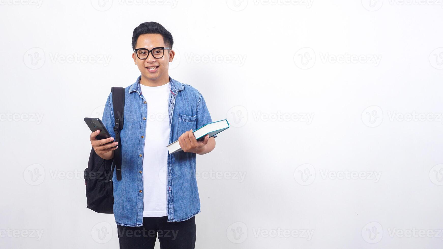 Handsome young Asian male university student in casual outfit with backpack is holding book and smartphone isolated on white background photo