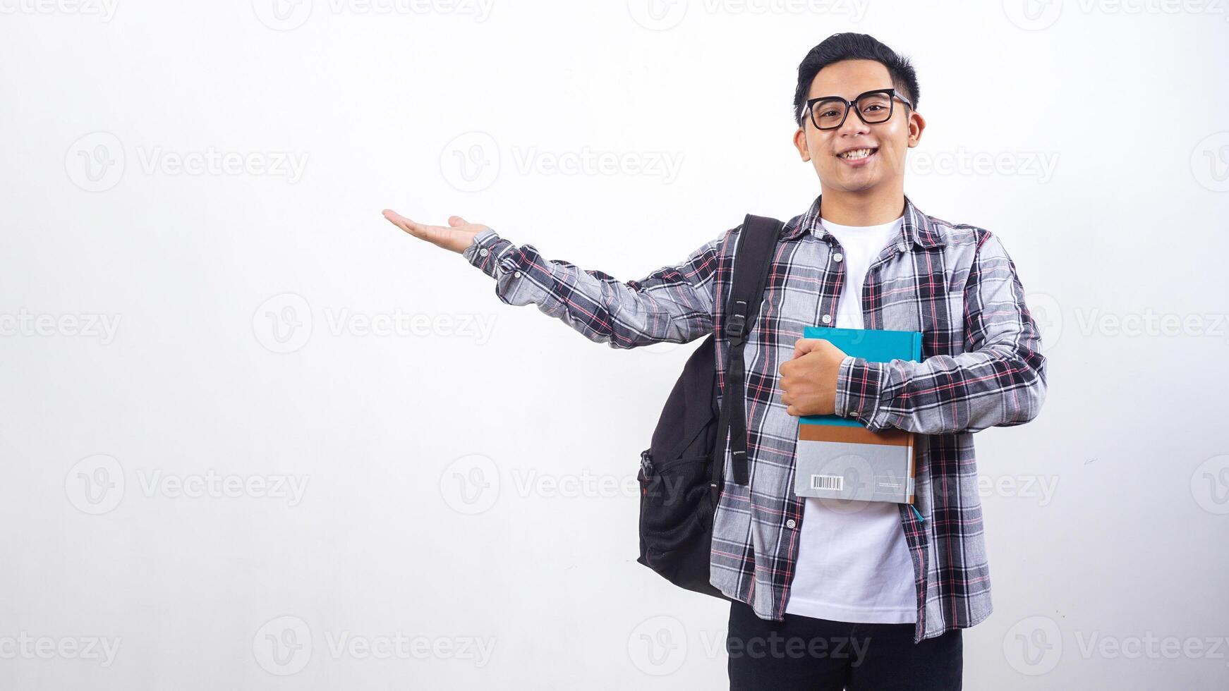 Smiling young Asian male college student with a backpack holding books in front of his chest and presenting empty copy space place isolated on white background photo