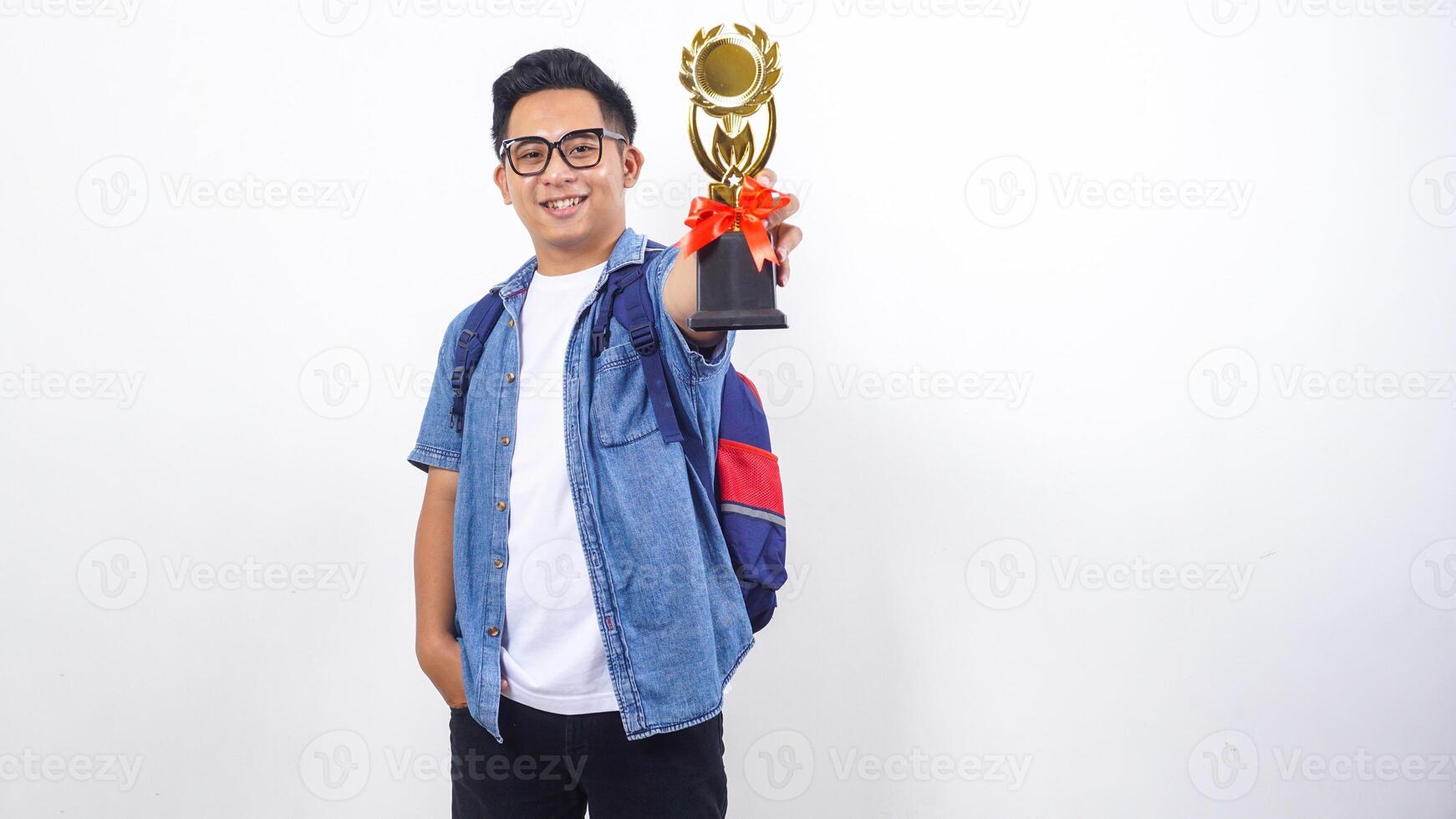 Smiling young Asian male university student in casual outfit with backpack is holding trophy isolated on white background photo