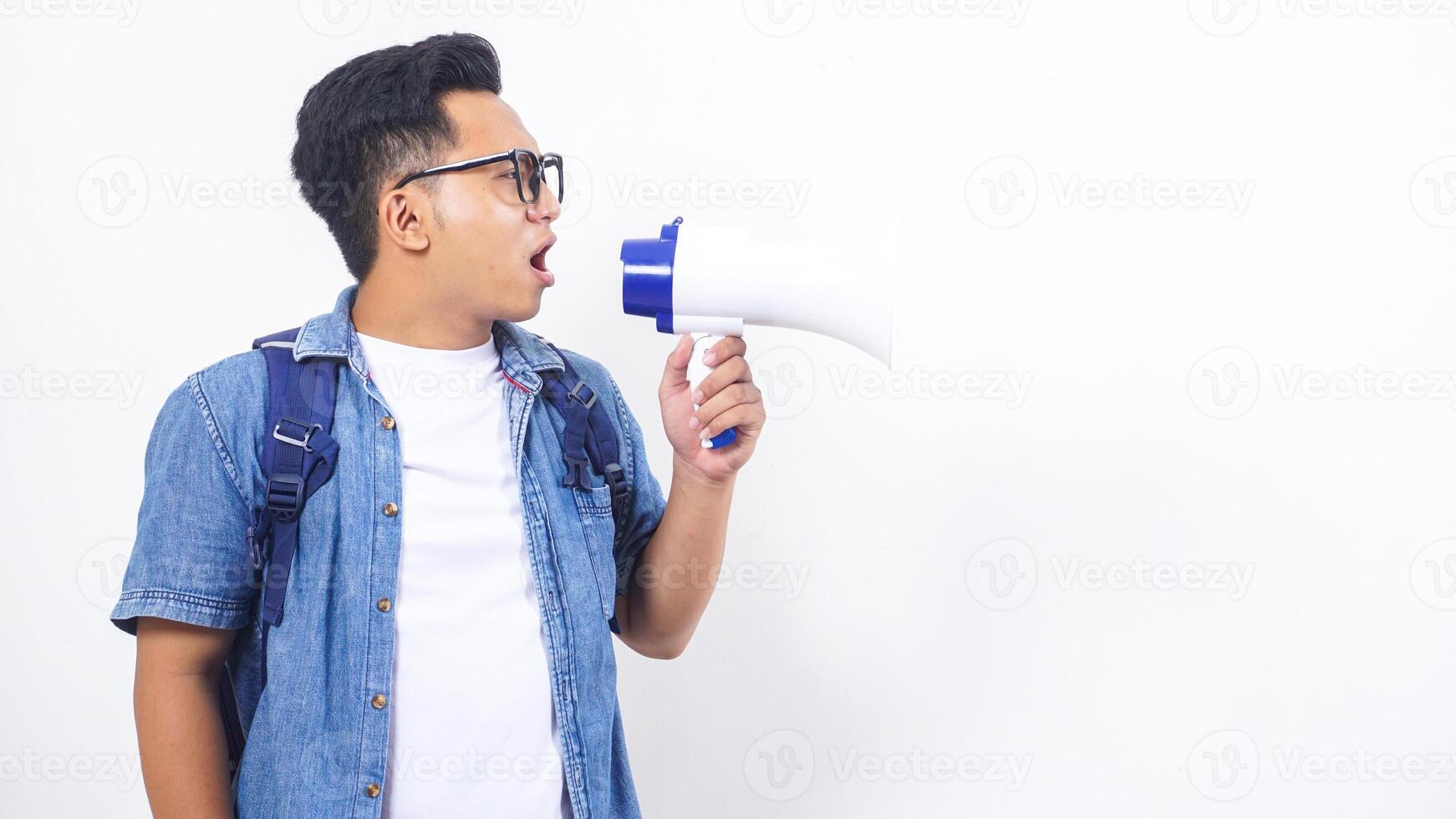 Portrait of excited young Asian male university student in casual outfit with backpack is shouting on megaphone isolated on white background photo