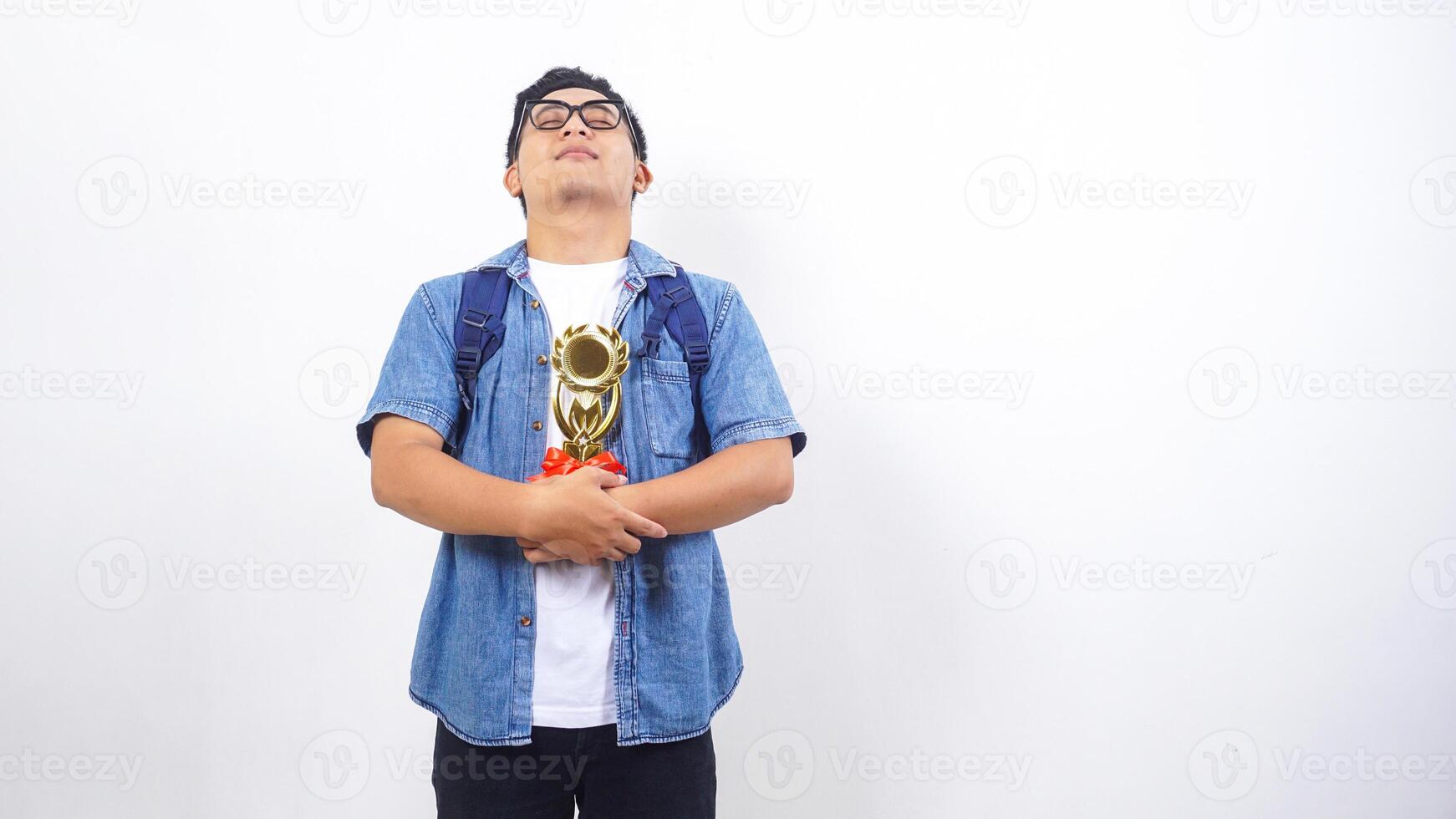 Young Asian male university student in casual outfit with backpack is holding trophy isolated on white background with copy space photo