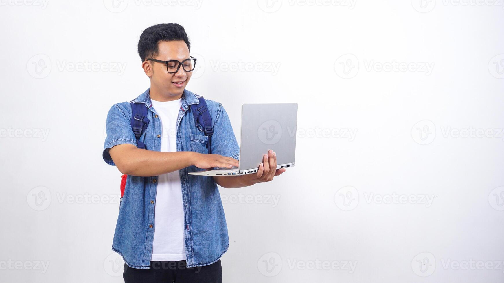 Smiling young Asian male university student in casual outfit with backpack and open laptop isolated on white background with copy space photo