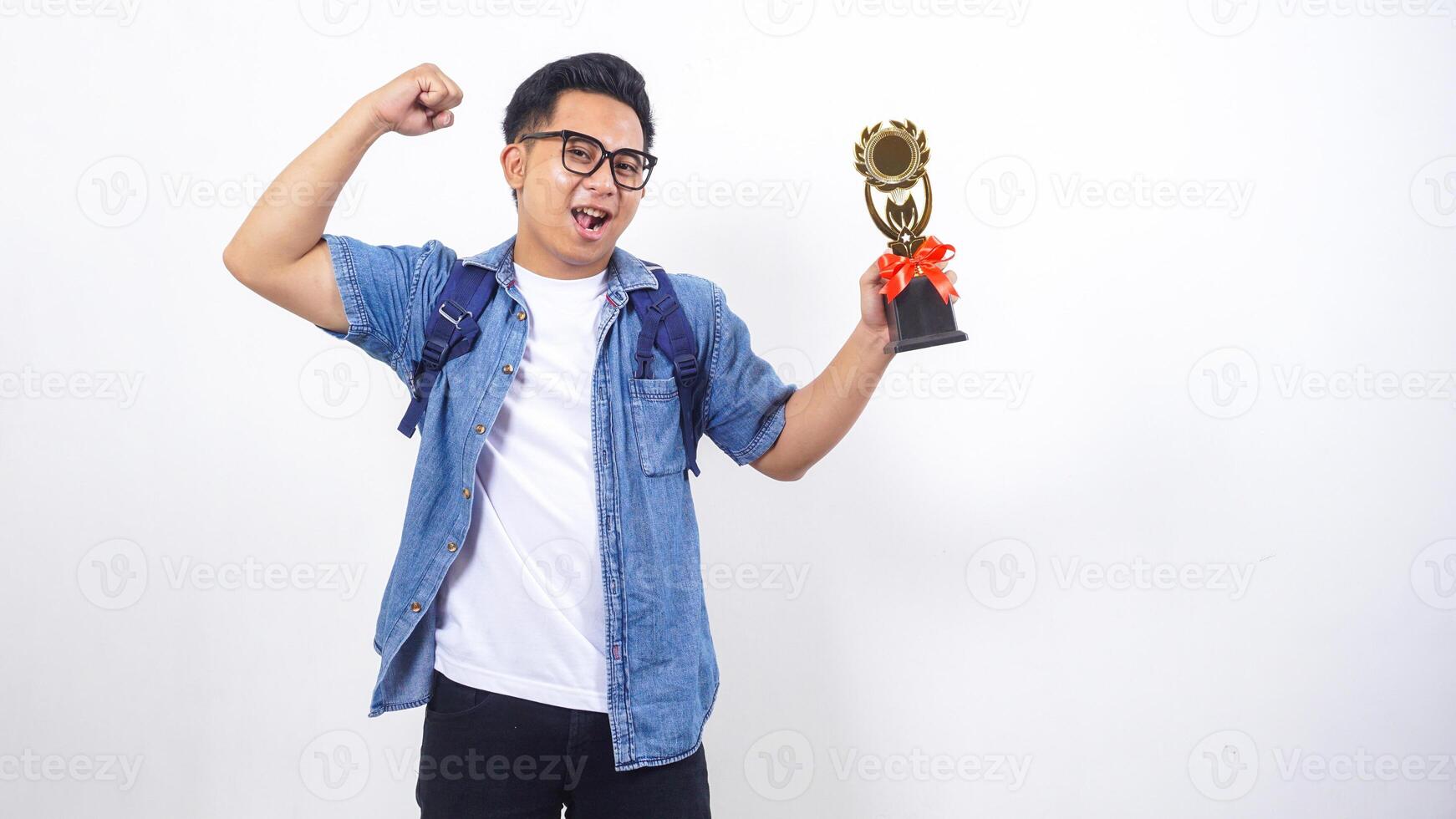 Happy young Asian male university student in casual outfit with backpack is holding trophy isolated on white background photo