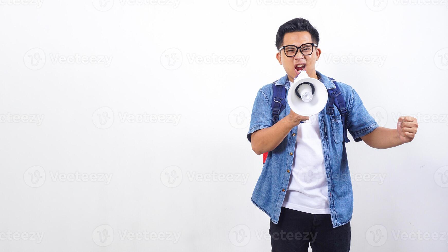 Portrait of unhappy young Asian male university student in casual outfit with backpack is shouting on megaphone isolated on white background with copy space photo