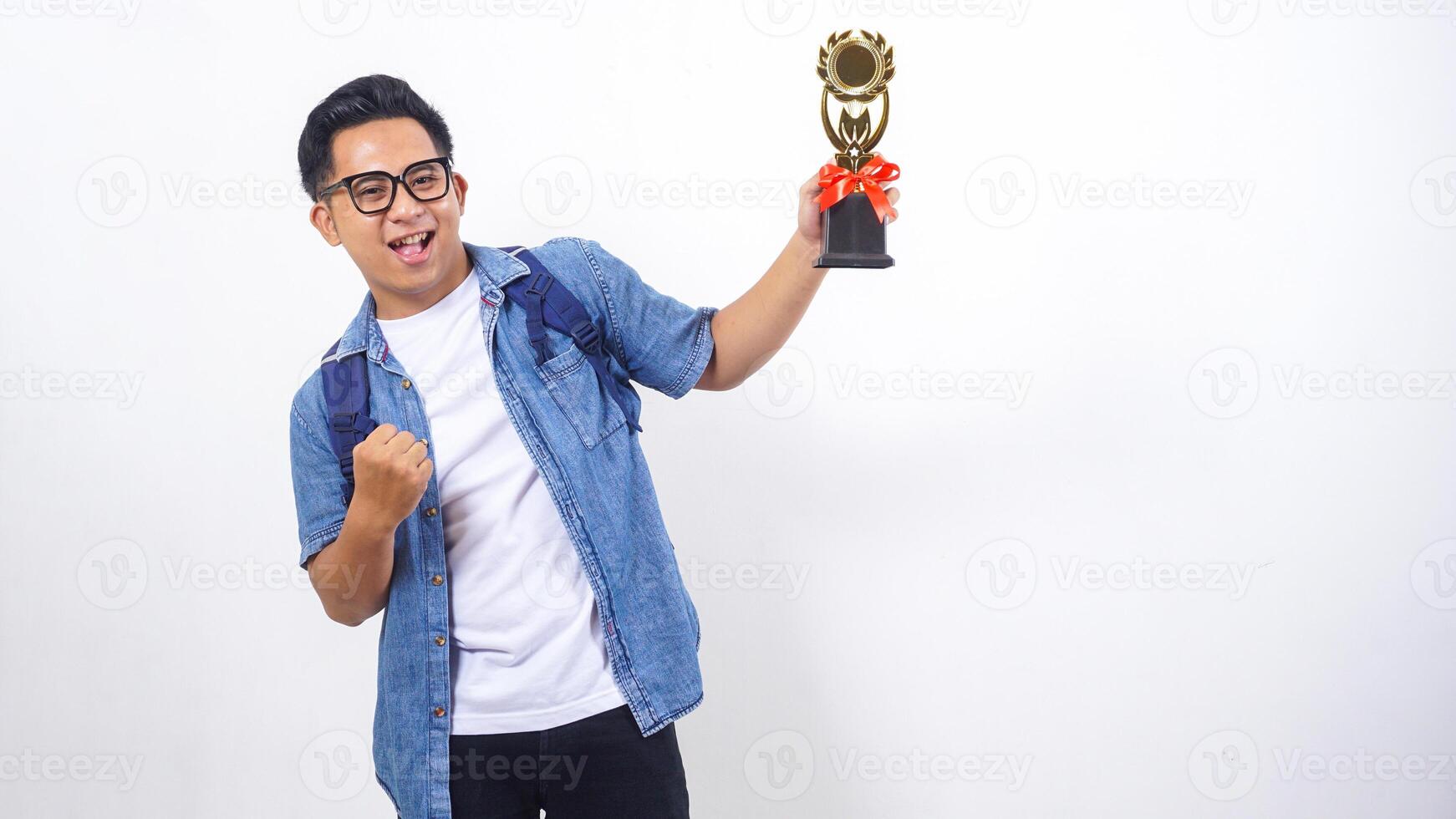 Excited young Asian male university student in casual outfit with backpack is holding trophy isolated on white background photo