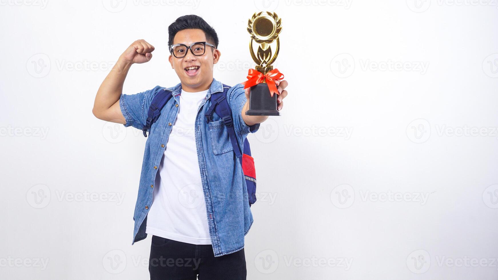 Excited young Asian male university student in casual outfit with backpack is holding trophy isolated on white background with copy space photo
