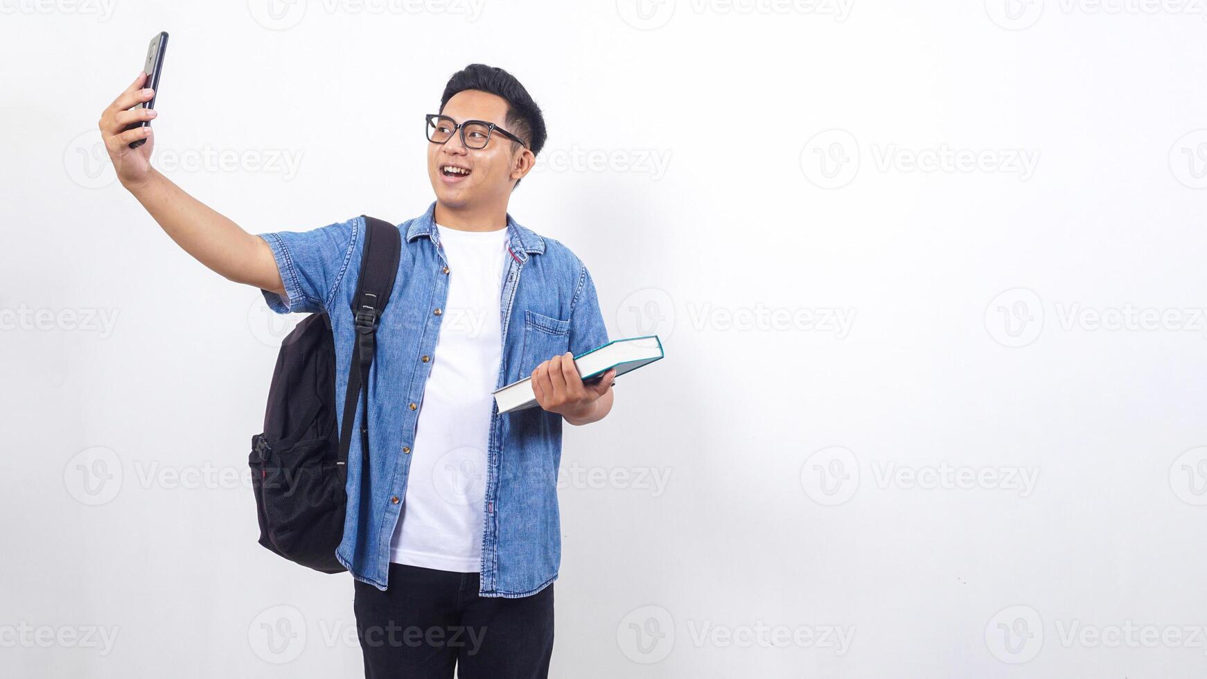 Handsome young Asian male university student in casual outfit with backpack is holding book and smartphone isolated on white background with copy space photo