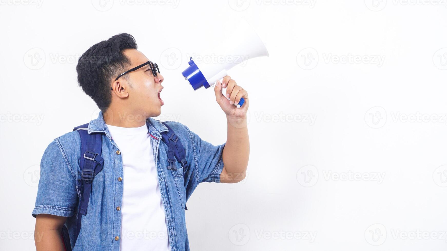 Portrait of unhappy young Asian male university student in casual outfit with backpack is shouting on megaphone isolated on white background photo