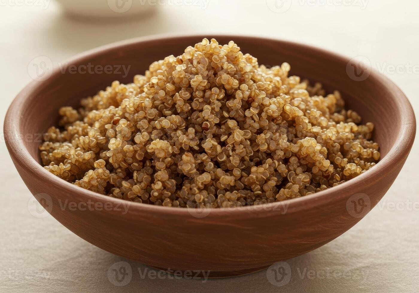 A brown bowl of cooked quinoa sits invitingly on a textured, neutral colored surface. photo