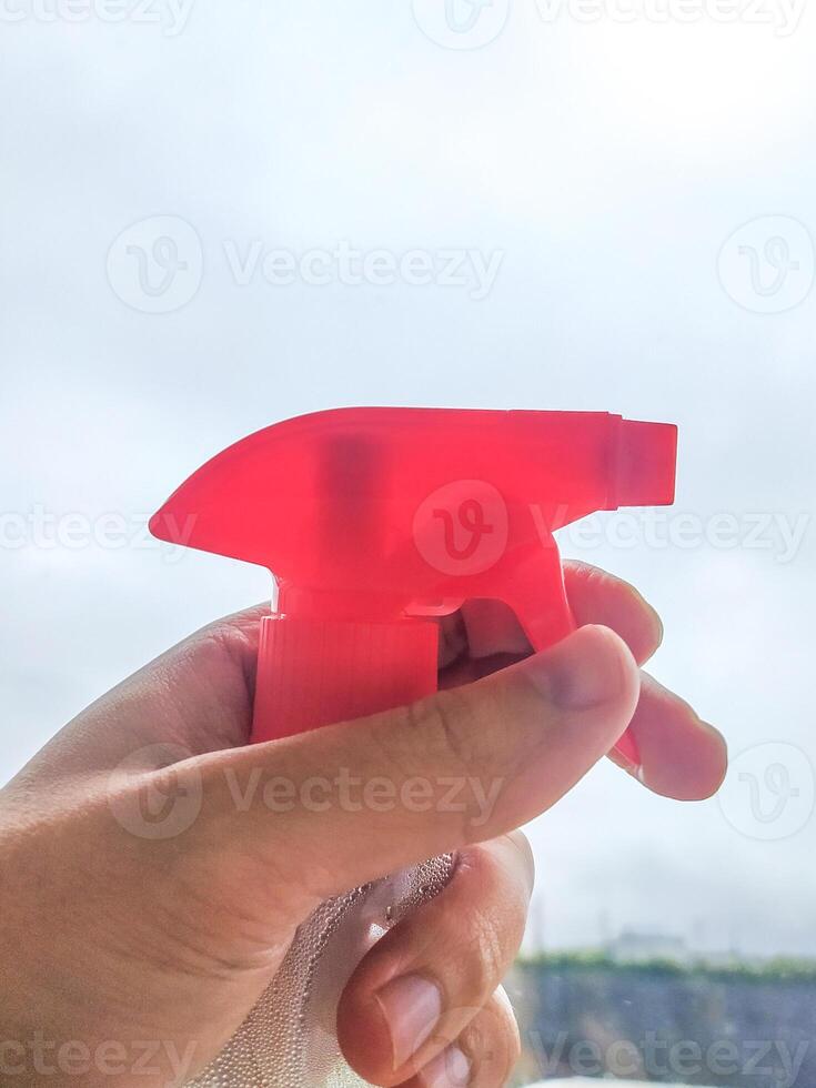 Hand holding a spray bottle with a vibrant red nozzle against a bright, overcast sky. Illustrates cleaning, hygiene, and household tasks. Useful for cleanliness or freshness concepts. photo