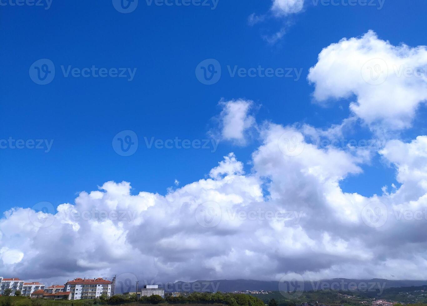 Vibrant skyscape featuring a clear blue sky contrasted with dramatic, fluffy white clouds. A cityscape faintly visible at the horizon. Evokes peace and wonder. Ideal for travel. photo