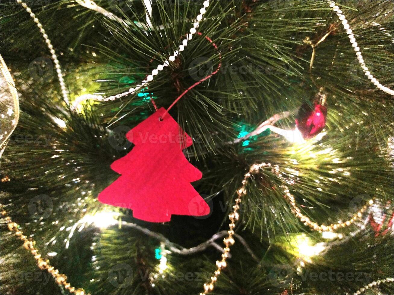 Close-up of a Christmas tree adorned with twinkling lights, red ornament, and elegant gold and pearl garlands. A charming red felt tree-shaped decoration adds a festive touch. photo