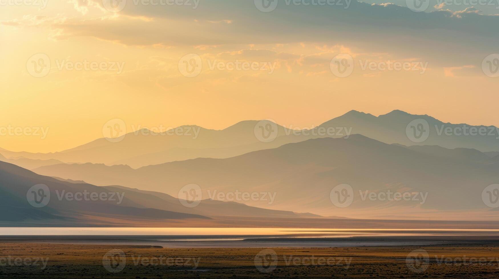 Layered mountain range fading into a hazy, golden sunset or sunrise sky. Still water reflects the light in a vast, tranquil landscape photo