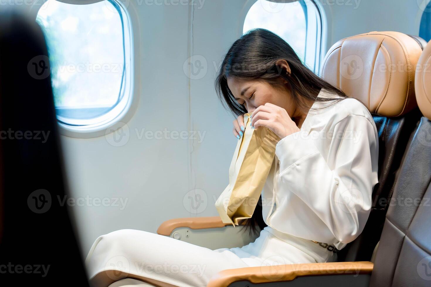 Young woman feeling air sickness on airplane, using vomit bag during flight, experiencing nausea and discomfort while seated in passenger cabin photo