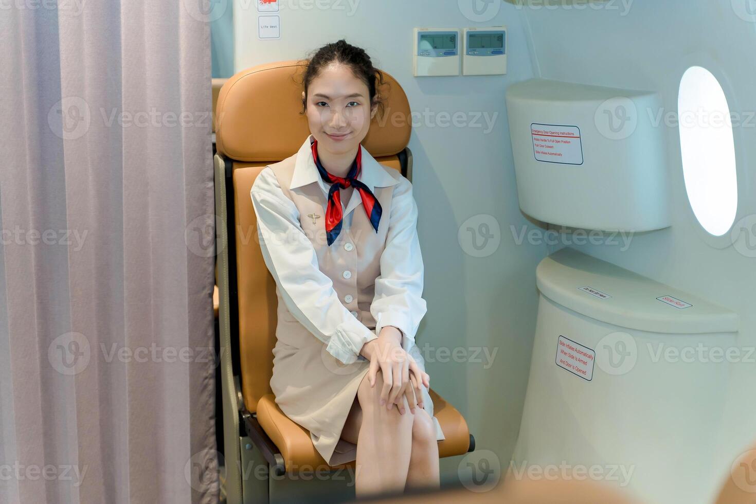Smiling flight attendant seated on jumpseat with hands folded, ready for takeoff procedures inside passenger aircraft, showing calm and professional demeanor photo