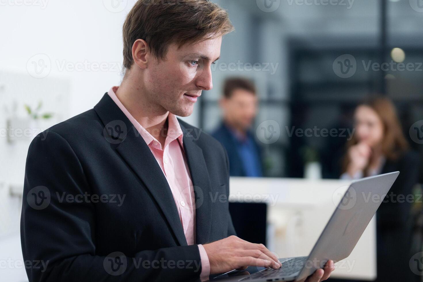 Professional man working on a laptop in a modern office setting with colleagues in the background during a productive meeting photo