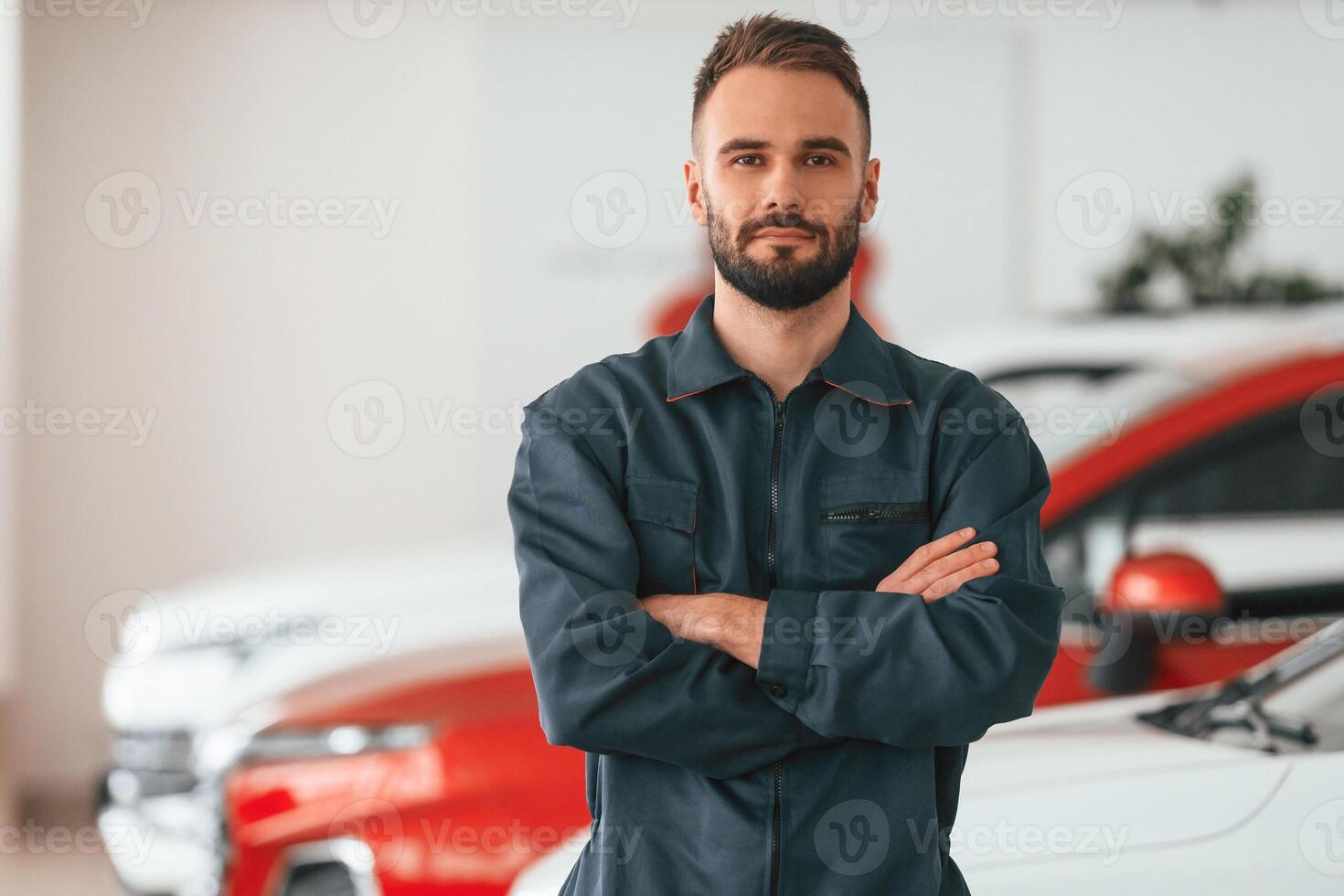With arms crossed, standing. Car repairman is in the garage with automobile photo
