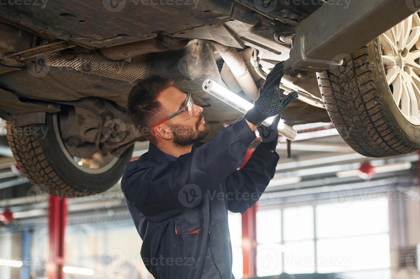 In blue colored uniform. Car repairman ,,is in the garage with automobile photo