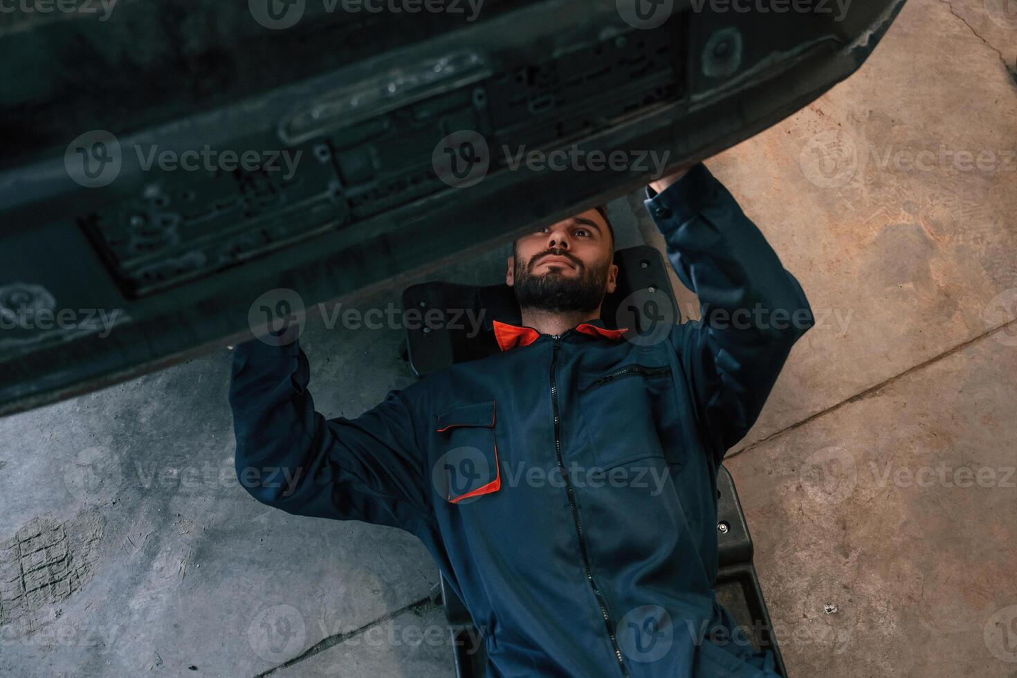 Lying down on the ground. Car repairman is in the garage with automobile photo