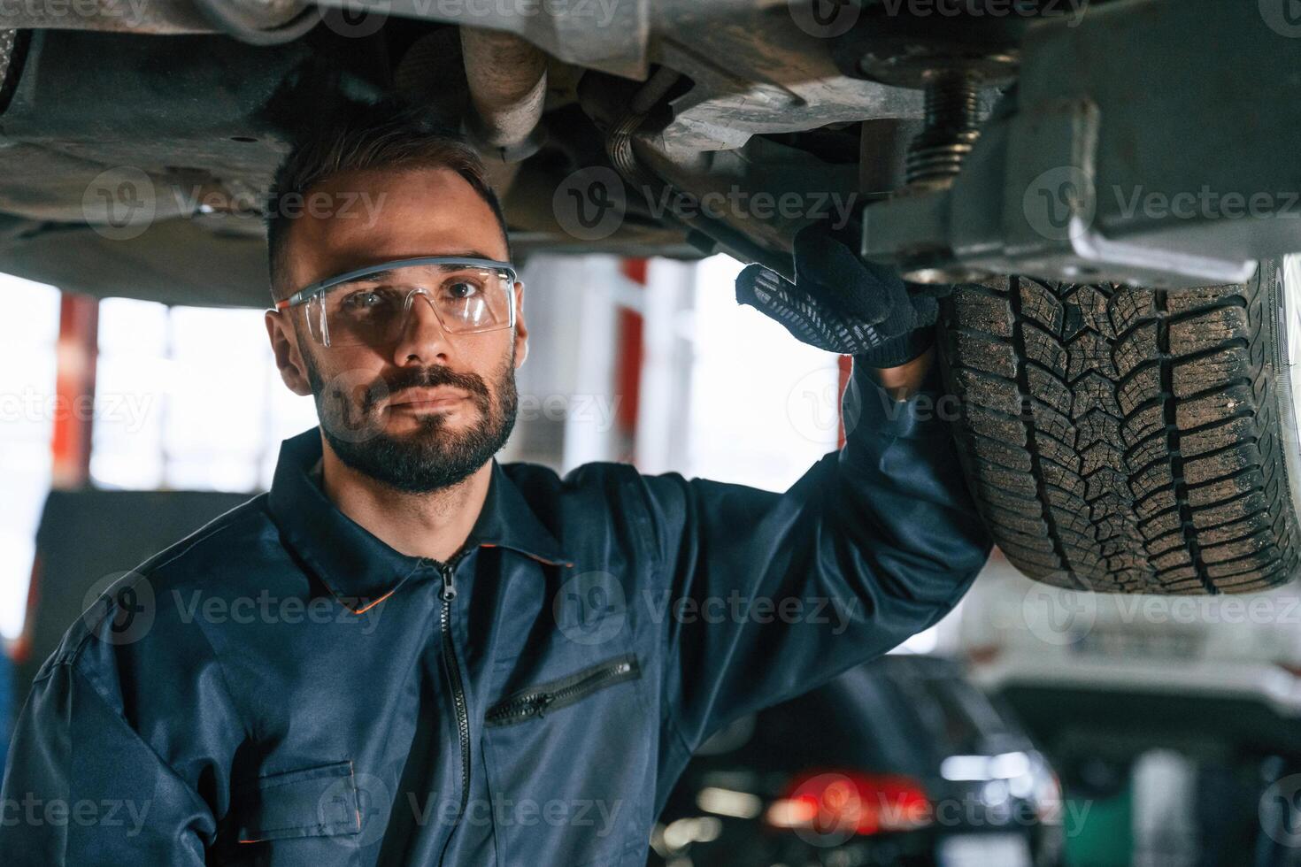 In protective eyewear, under vehicle,. Car repairman is in the garage with automobile photo