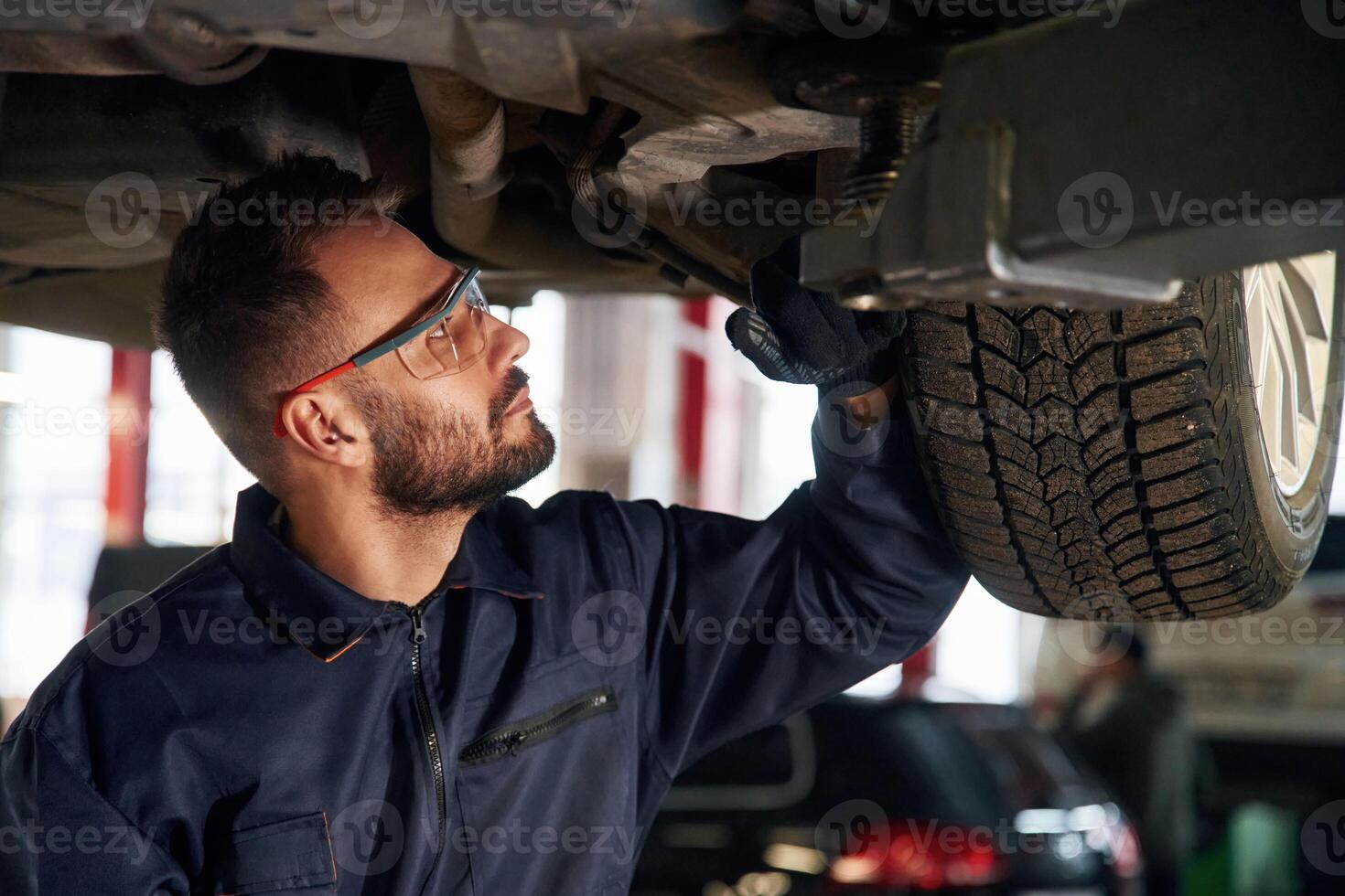 In protective, eyewear, under vehicle. Car repairman is in the garage with automobile photo