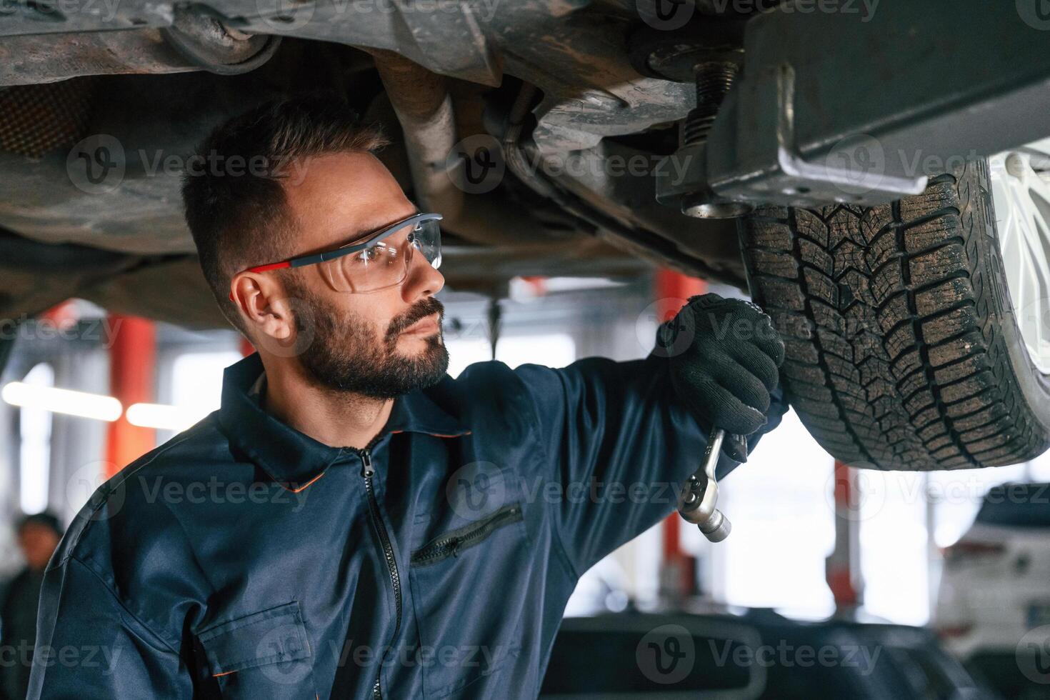 In protective eyewear, under ,vehicle. Car repairman is in the garage with automobile photo