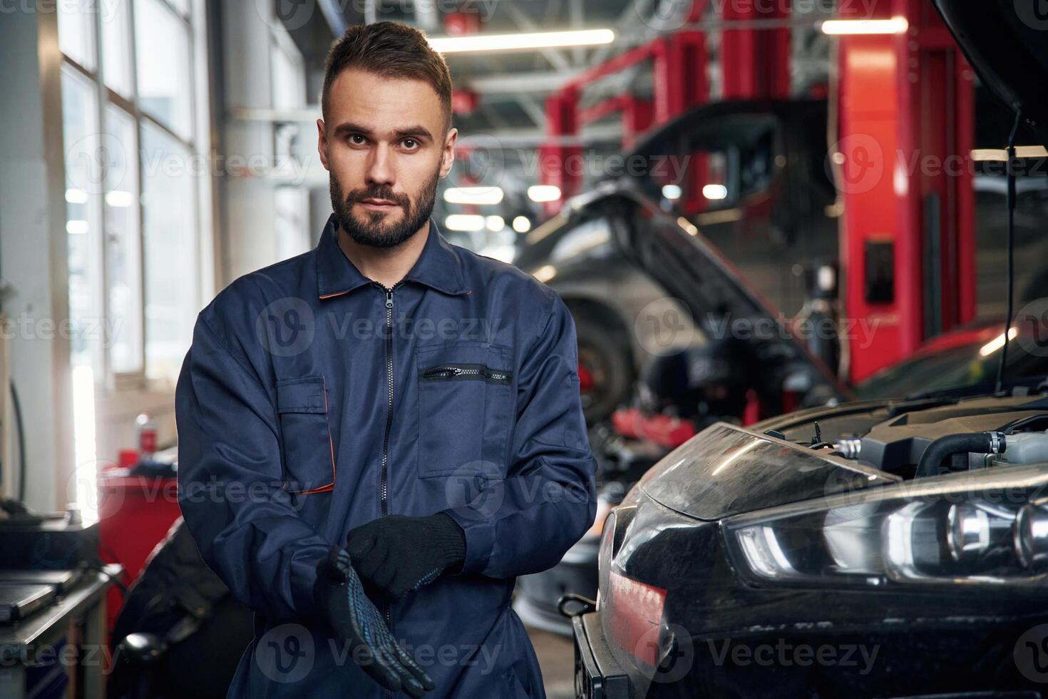 Wearing ,the ,gloves, standing. Car repairman is in the garage with automobile photo
