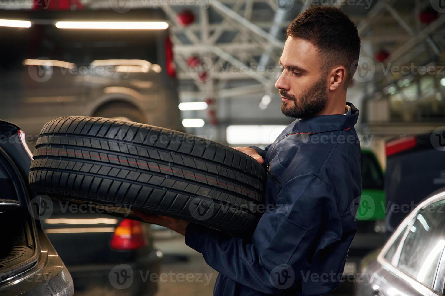 Holding ,a ,tire. .Car repairman is in the garage with automobile photo