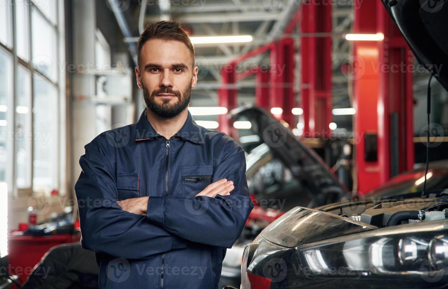 With arms crossed, standing.. Car repairman is in the garage with automobile photo